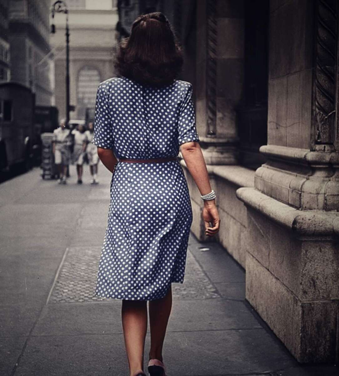 A woman in a blue polka dot dress walks down a city sidewalk, seen from behind. She has shoulder-length brown hair and wears a bracelet. Tall buildings and other pedestrians line the street.