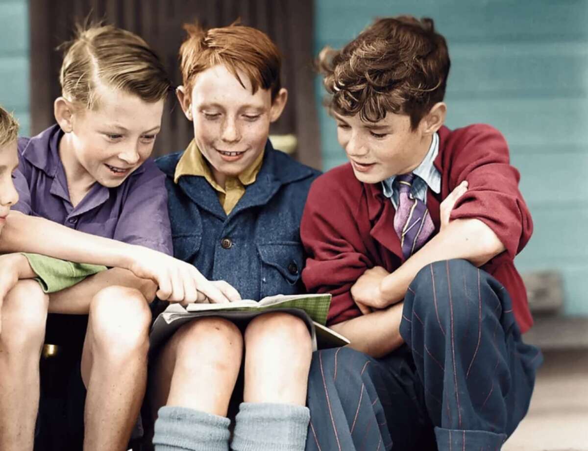 Three boys sit closely together, smiling and looking at a book or notebook on one boy’s lap. They appear engaged and happy, with one boy pointing at the page. The background is blurred but shows a blue wall.