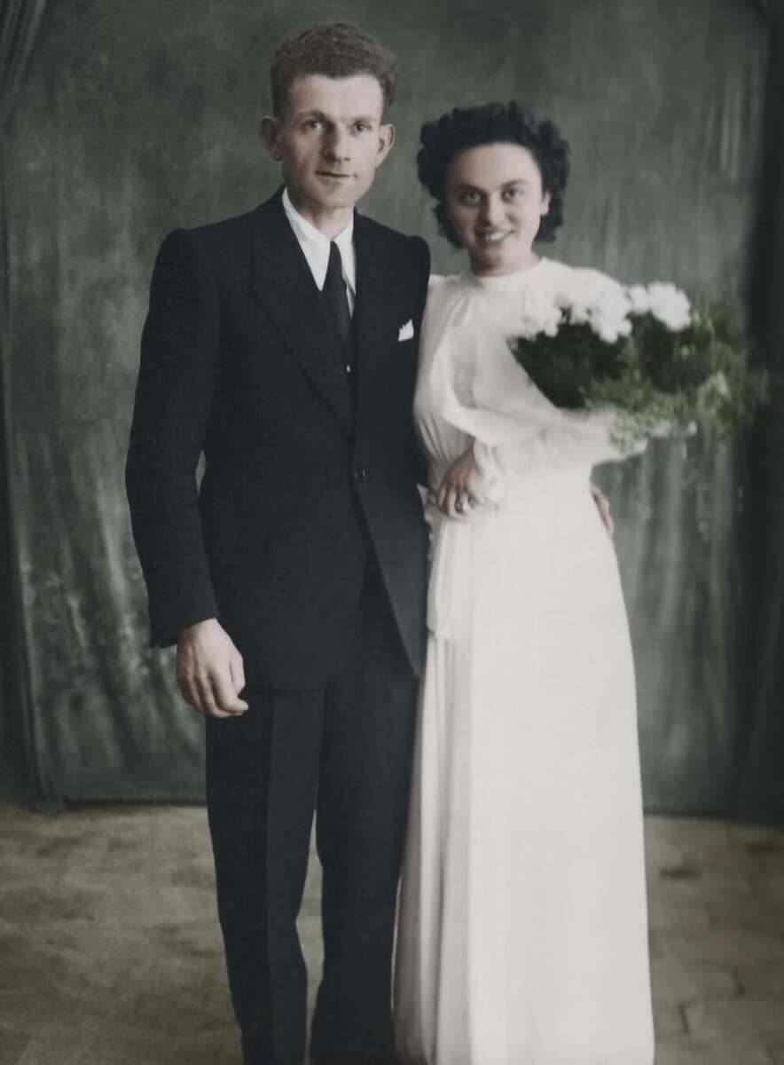 A vintage photo of a bride and groom poses together. The groom wears a black suit and tie; the bride wears a white dress and holds a bouquet of white flowers. They stand in front of a simple, dark curtain backdrop.
