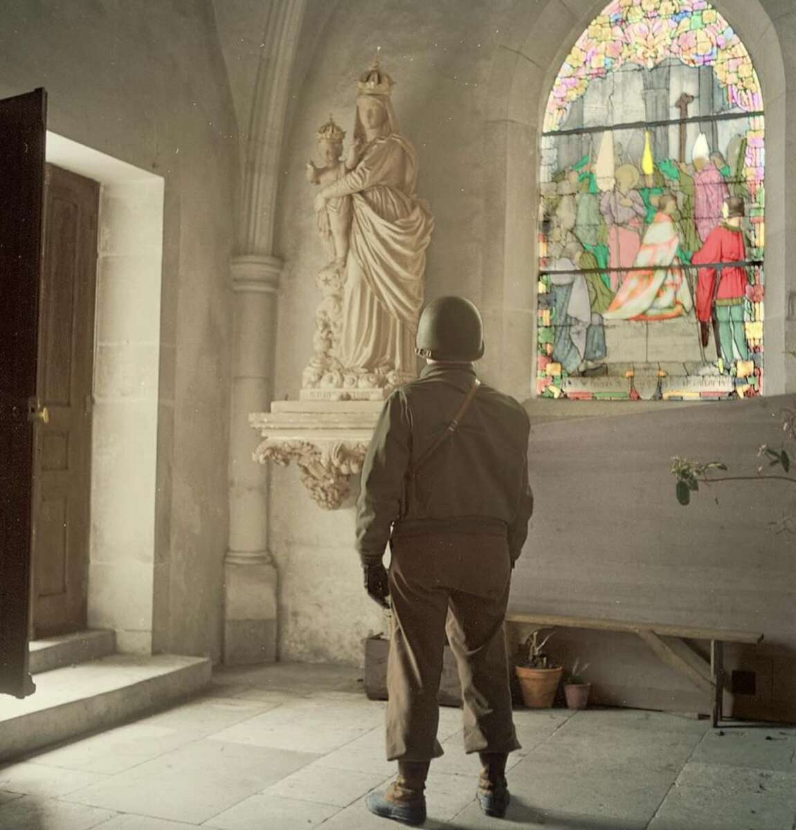 A soldier in uniform and helmet stands inside a church, facing a statue of the Virgin Mary holding baby Jesus. Sunlight streams through a colorful stained glass window next to the statue.