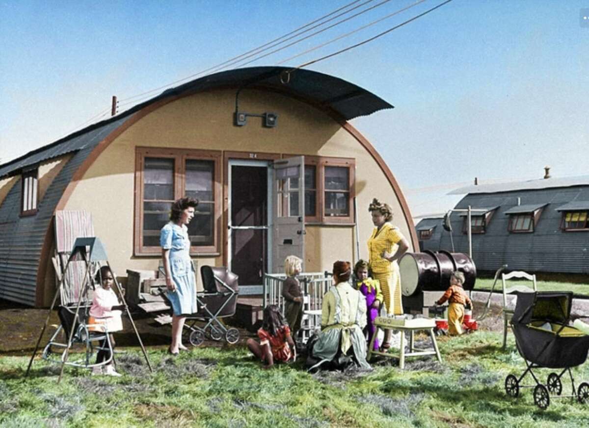 A group of women and children gather outside a Quonset hut on grass, surrounded by toys, chairs, and a stroller, with another identical hut and metal barrels in the background under a clear sky.
