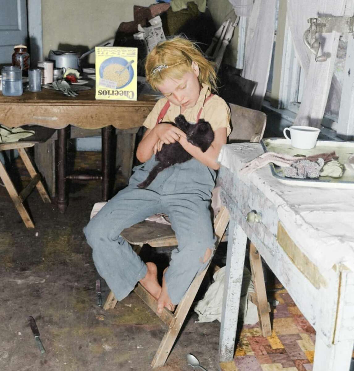 A young girl with blonde hair sits barefoot on a wooden chair in a cluttered room, gently holding a black kitten. She wears blue overalls and a yellow shirt. The room has worn furniture, dishes, and a cereal box on the table.