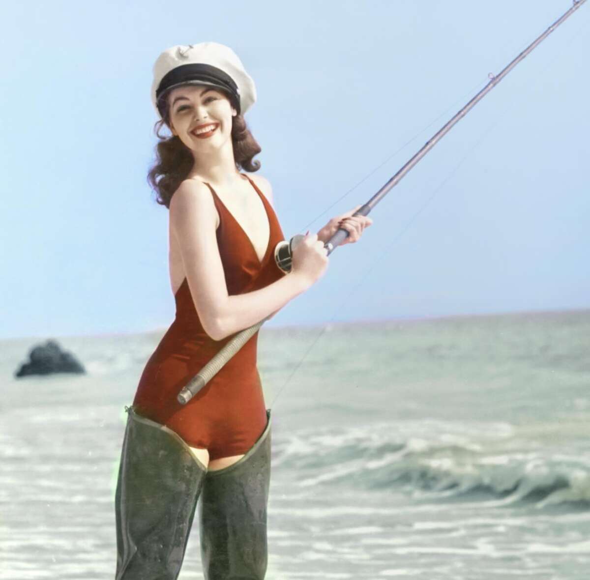 A smiling woman in a red swimsuit, white sailor hat, and waders stands in shallow ocean water, holding a fishing rod with gentle waves and a blue sky in the background.