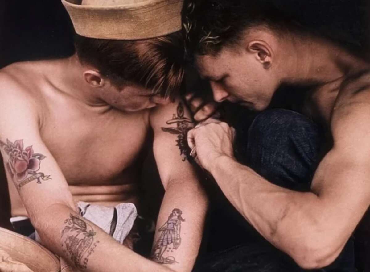Two shirtless men sit closely together. One, wearing a sailor hat, looks down at his arm while the other man tattoos a design onto it. Both have visible tattoos on their arms.