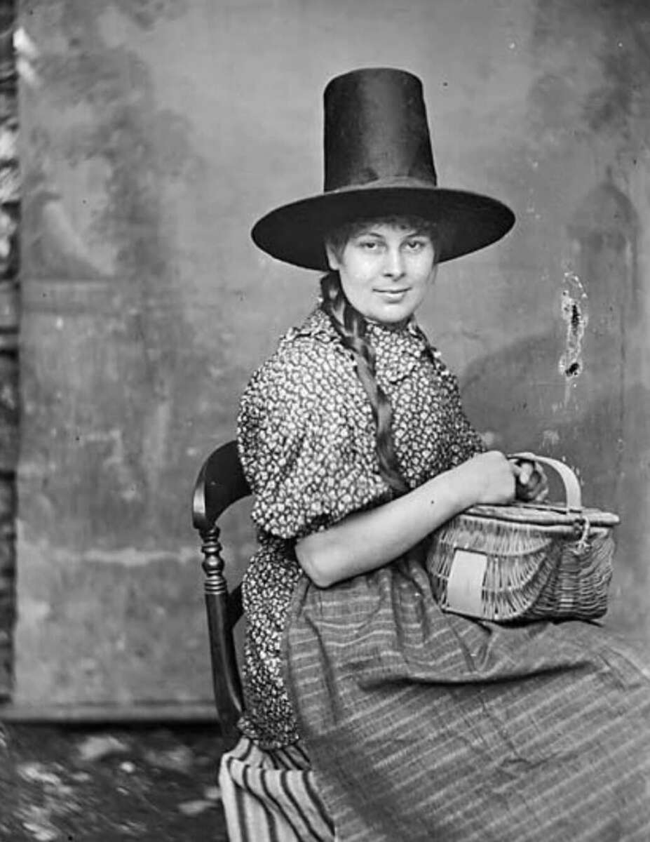 A young woman in a tall, wide-brimmed hat and patterned dress sits on a chair, holding a woven basket in her lap. Her hair is in braids, and she looks at the camera with a slight smile. The photo is black and white.