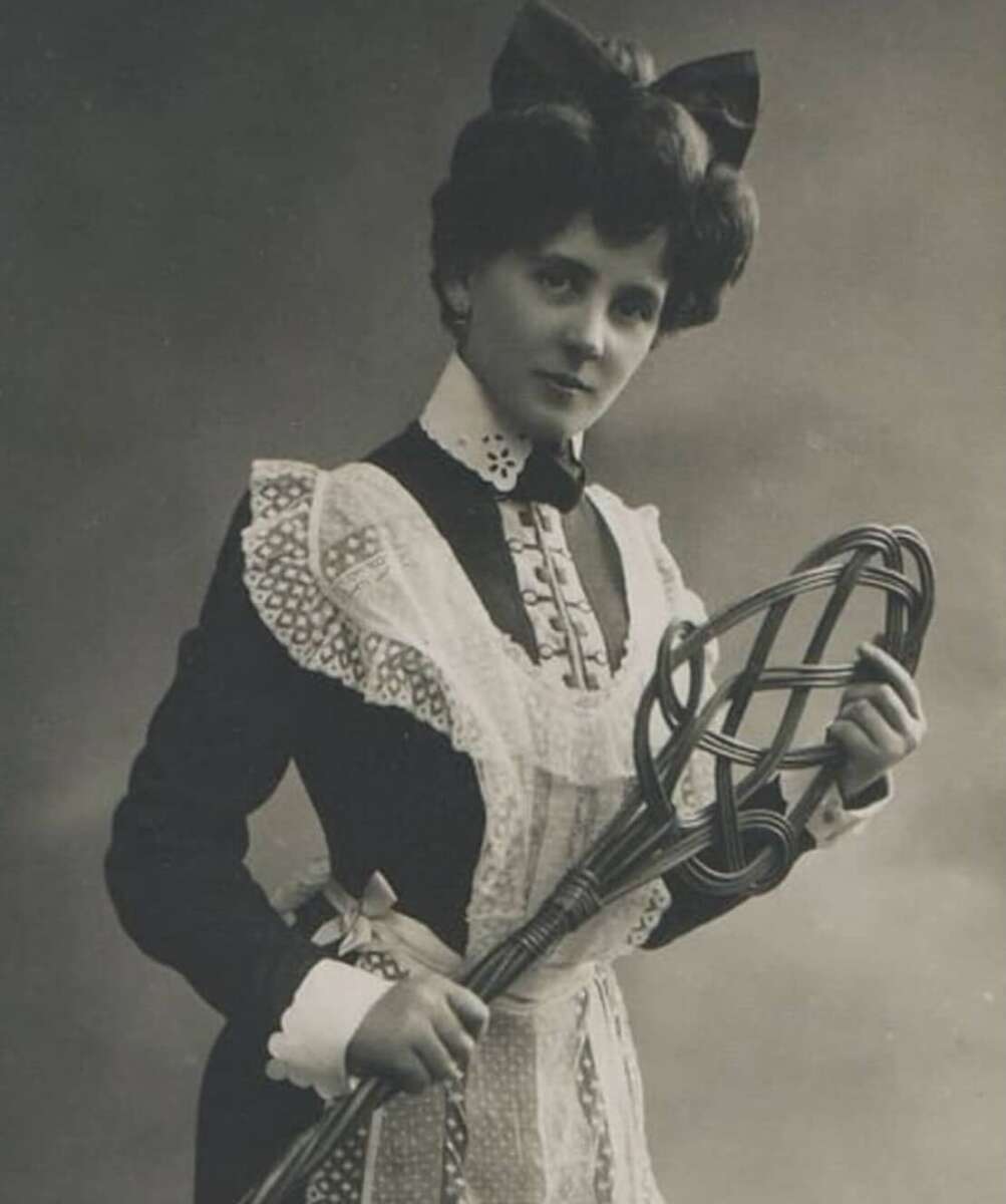 A woman in Victorian-era clothing with a lace apron and large bow poses holding an old-fashioned carpet beater, looking directly at the camera in a vintage black-and-white photo.
