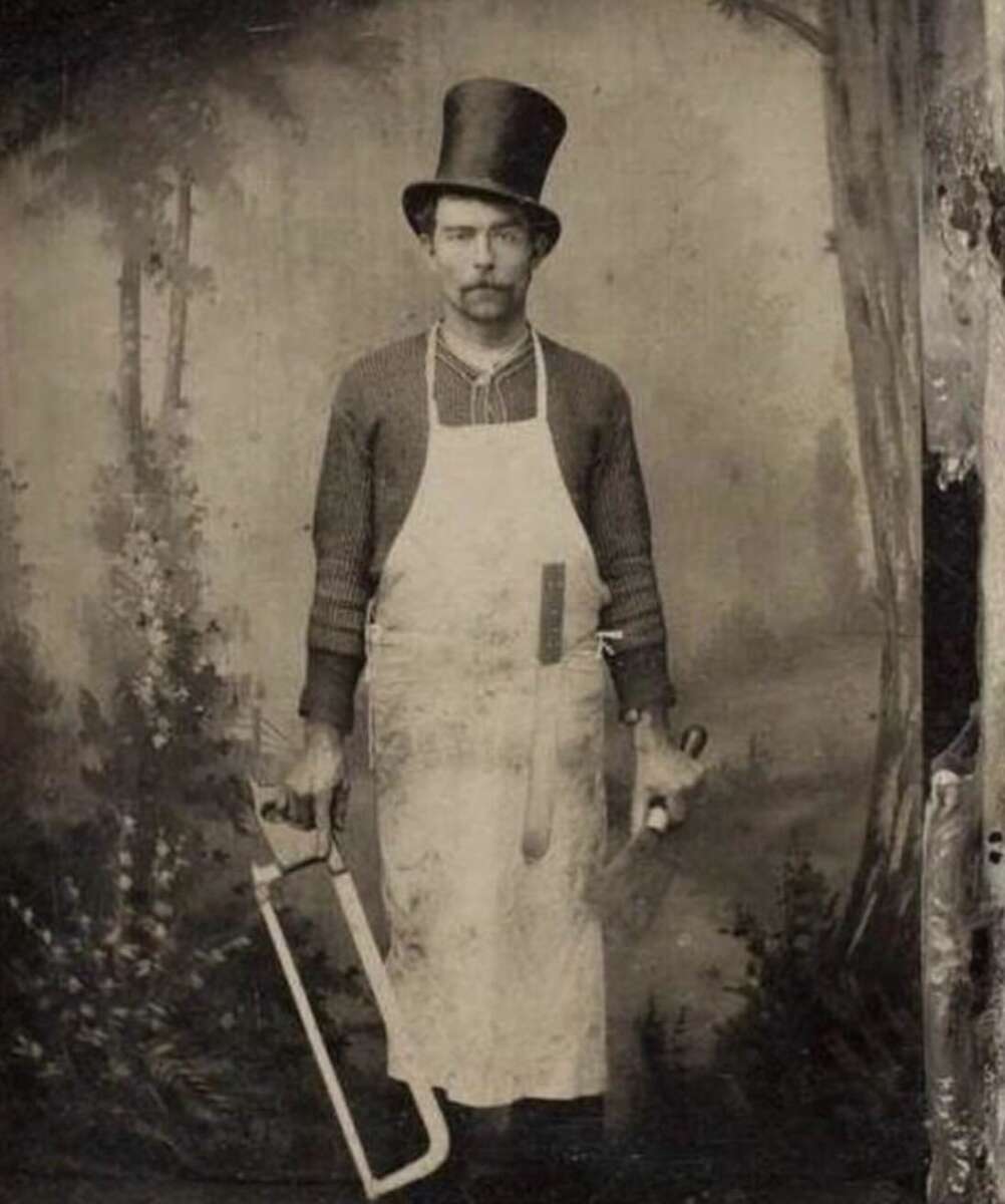 A vintage black-and-white photo of a stern man in a tall top hat and apron, holding a large saw and a knife, standing in front of a painted forest backdrop.