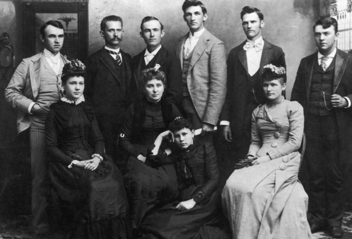 Black and white vintage photo of eleven young adults, six men in suits and five women in long dresses with floral headpieces, posed together indoors against a painted backdrop.