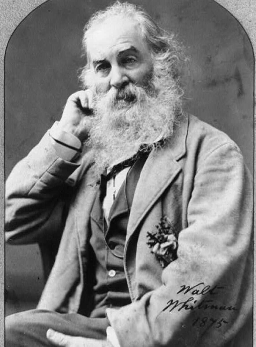 Black-and-white portrait of an older man with a long white beard and hair, wearing a suit, seated and resting his head on one hand. The photo is signed “Walt Whitman 1875” in the bottom right corner.