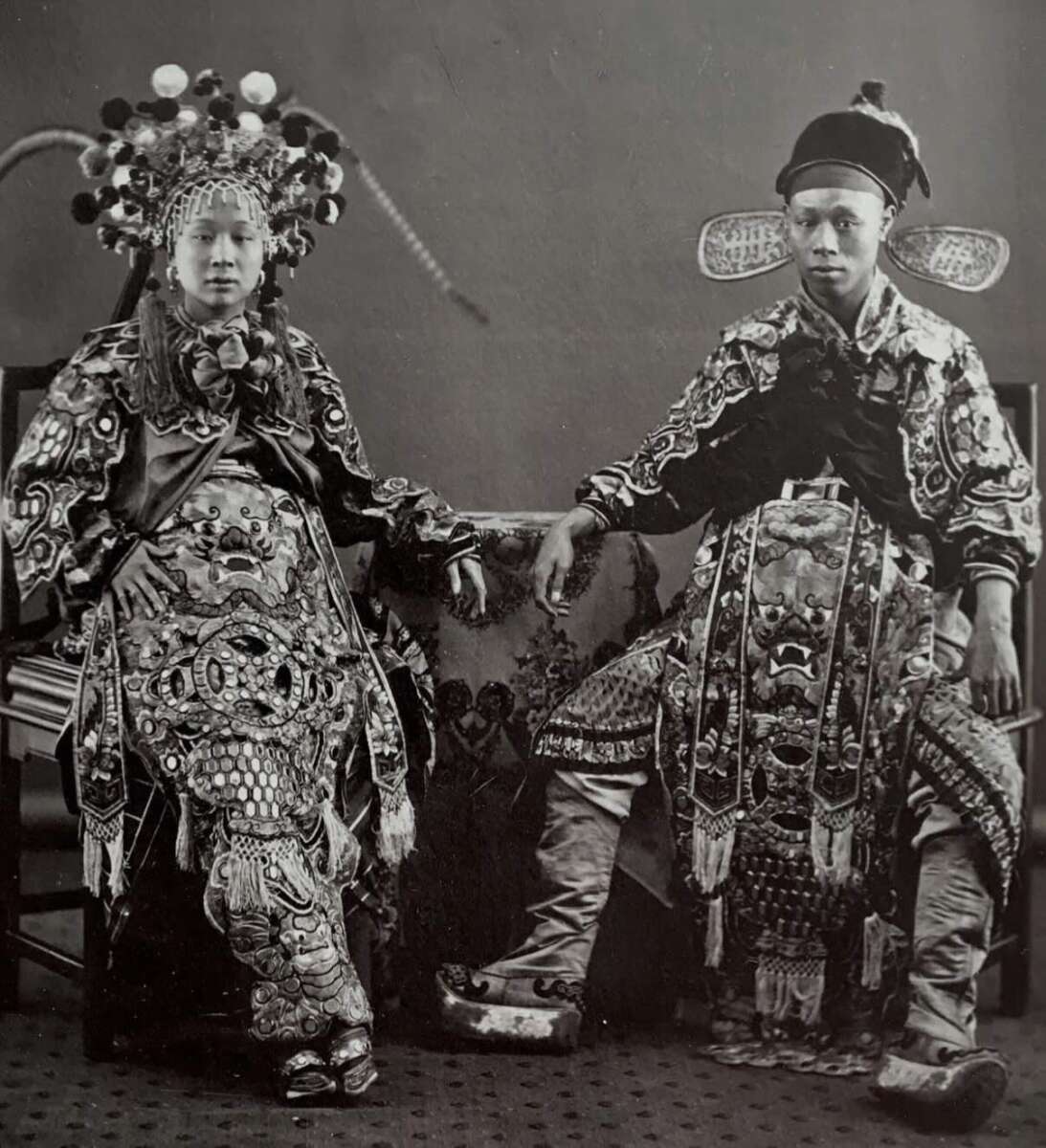 Two people dressed in elaborate traditional Chinese opera costumes sit side by side, posing for a black-and-white photograph. Their detailed garments feature ornate embroidery, headpieces, and patterned designs.