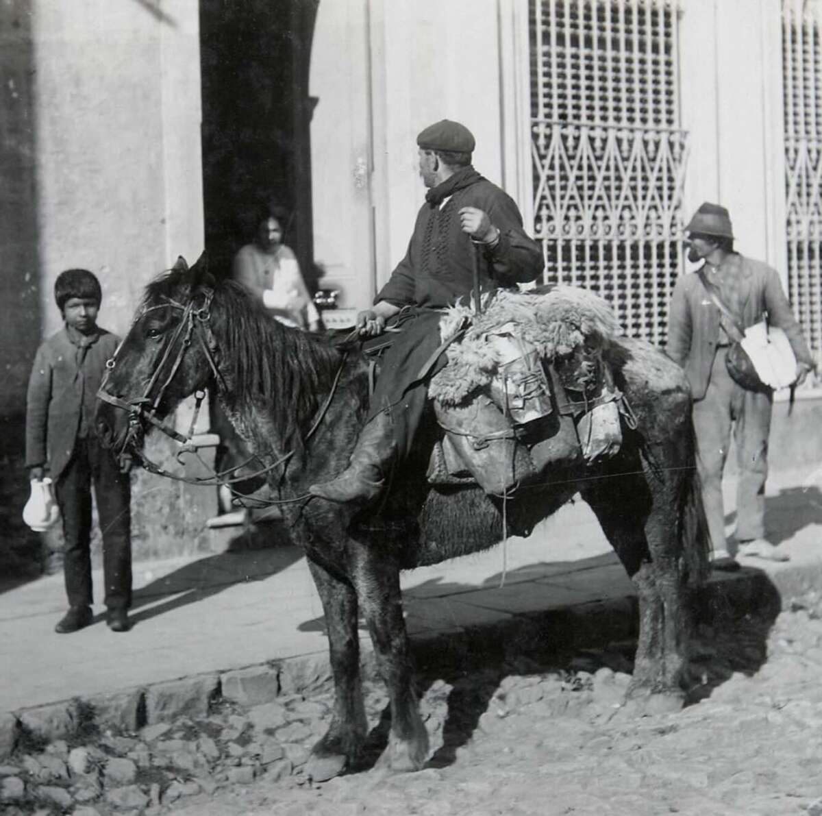 A man sits on a saddled horse on a cobblestone street, looking left. Three people stand nearby on the sidewalk in front of a building with barred windows. The scene appears to be from an earlier time period.
