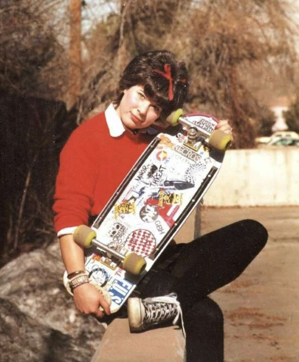 A person with short dark hair, wearing a red sweater, black pants, and black Converse sneakers, sits on a ledge outdoors holding a skateboard covered in colorful stickers.