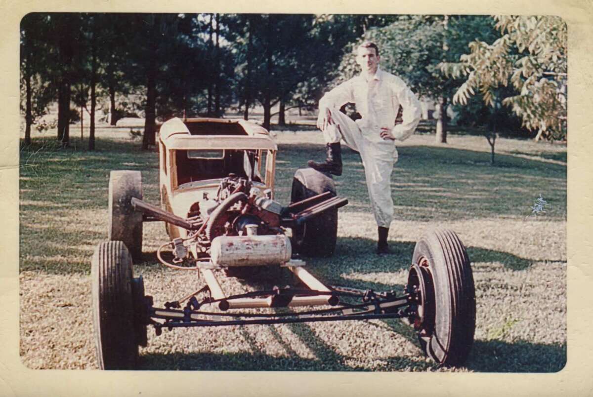 A man in a white jumpsuit stands outdoors with one foot on the front bumper of a partially assembled vintage car, surrounded by grass and trees.