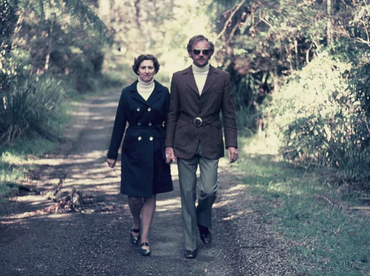 A man and woman walk hand in hand down a sunlit, tree-lined path. Both wear stylish 1970s outfits; the woman in a dark coat and the man in sunglasses, a brown blazer, white turtleneck, and light trousers.