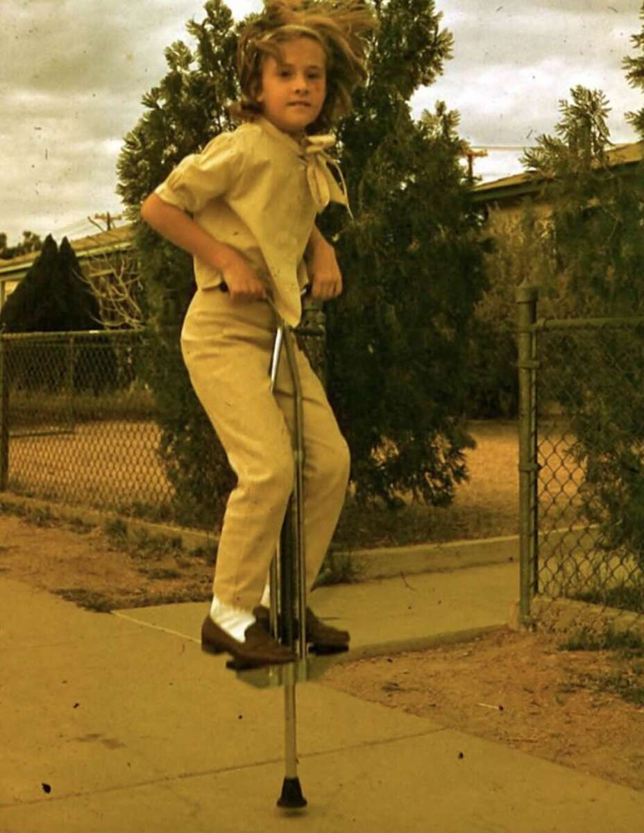A young person with shoulder-length hair and light-colored clothes is using a pogo stick on a sidewalk, with trees, a fence, and a house in the background. Their hair is flying up as they bounce.