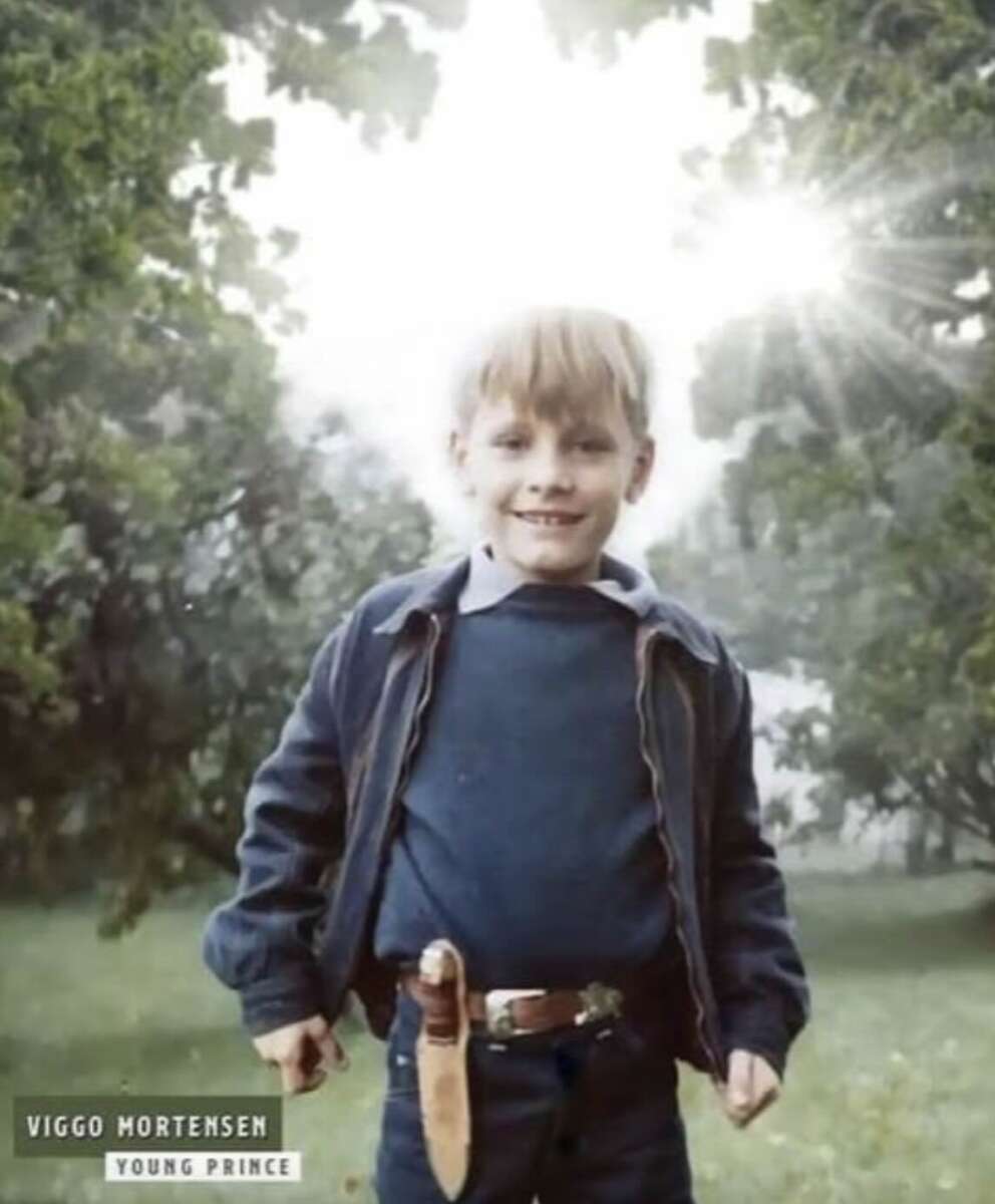 A young boy stands outdoors in a grassy area, smiling at the camera. He wears a blue jacket, blue shirt, and jeans, with a toy sword on his belt. Sunlight shines through the trees behind him. Text reads “Viggo Mortensen Young Prince.”