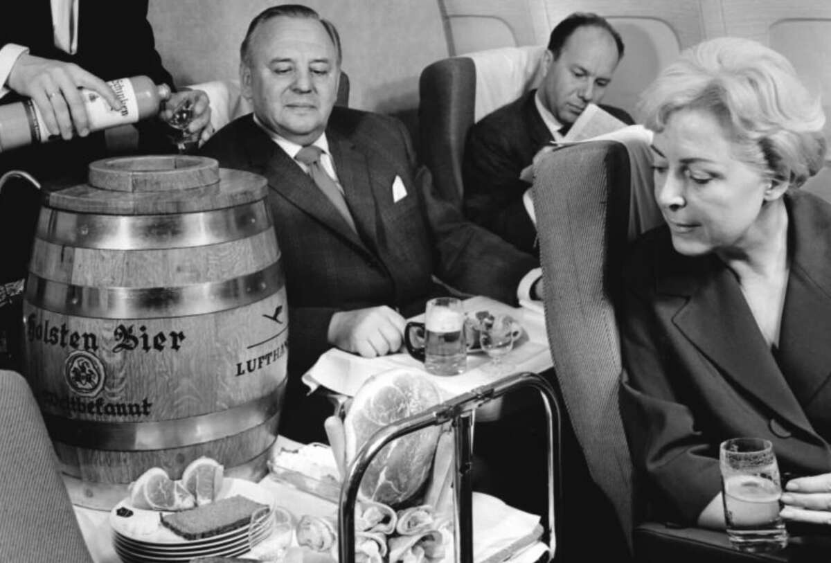 Passengers on an airplane are served beer from a large Holsten Bier keg, with glasses, food, and citrus slices on a tray. A woman in front looks to the side while two men sit behind her, one writing and one watching the beer being poured.