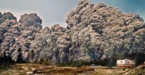 Massive, dark grey volcanic ash clouds billow into the sky behind a forested area and a white building, creating a dramatic scene of a powerful volcanic eruption.