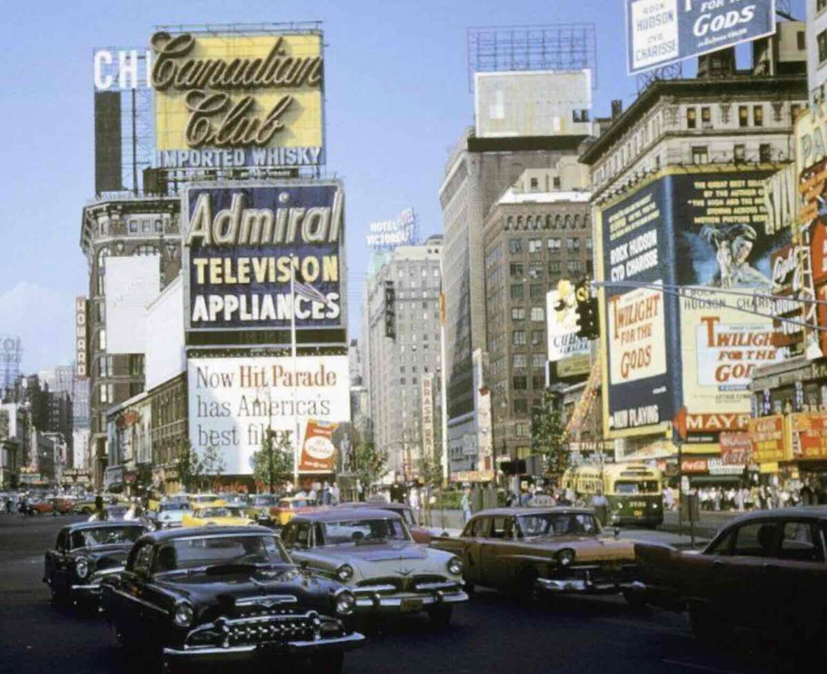 1950s Times Square scene with vintage cars, large colorful billboards advertising whisky, televisions, and films, and tall buildings in the background under a blue sky.