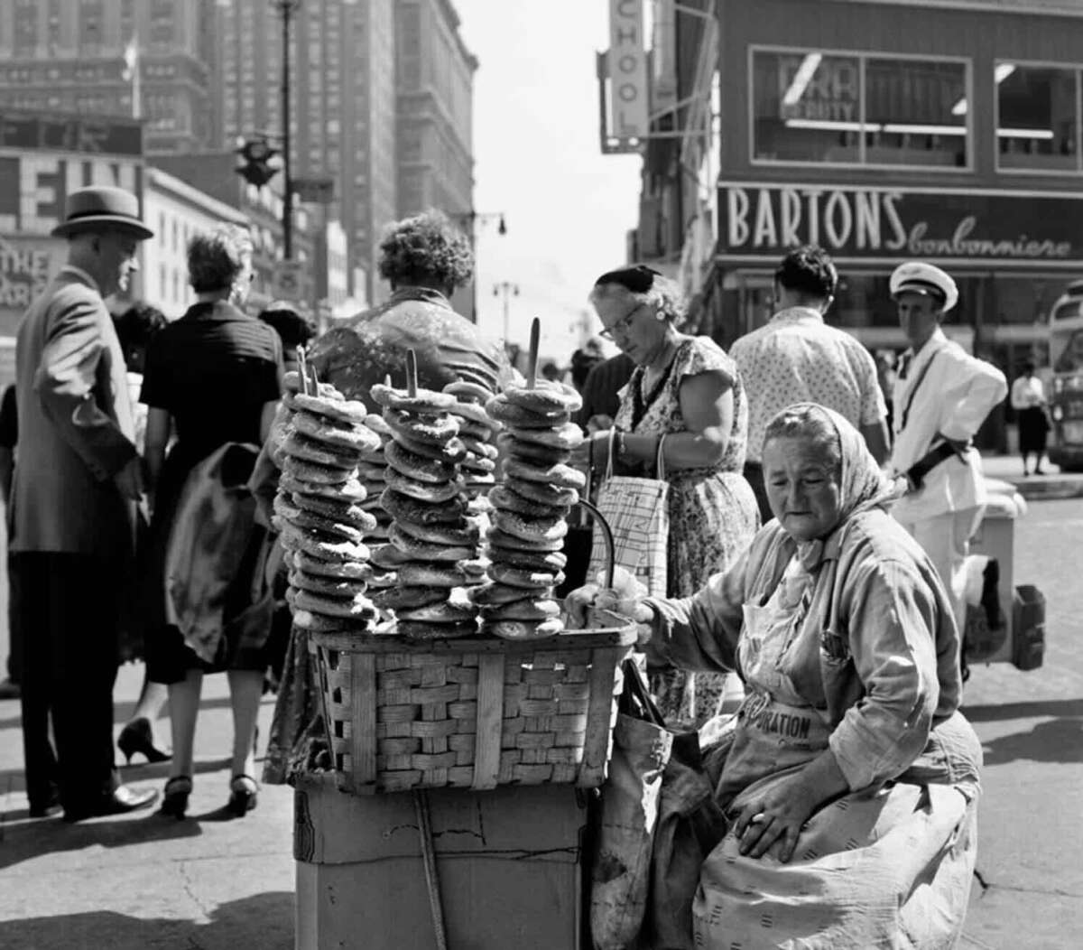 An elderly woman sits next to a large basket stacked with pretzels on skewers, selling them on a busy city street. Pedestrians walk by, and storefronts and signs are visible in the background.