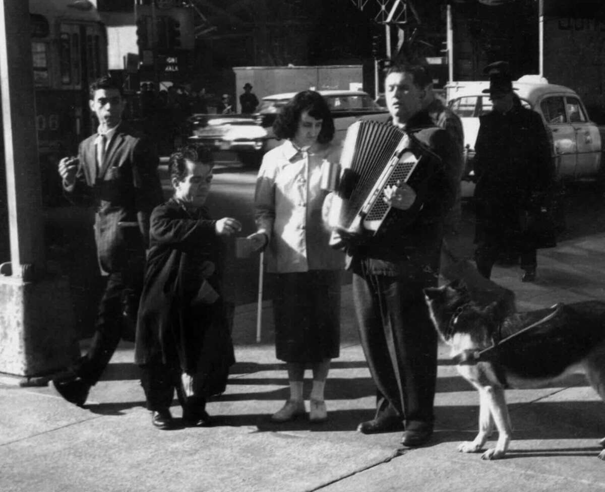 A street scene in black and white shows a man playing an accordion, a woman and two men standing nearby, and a German Shepherd dog on a leash; cars and pedestrians are in the background.