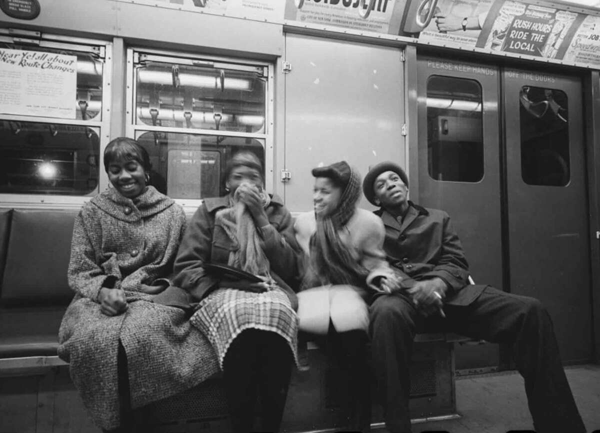 Four people sit closely together on a subway bench, laughing and smiling. They wear winter coats and hats, and the subway car is dimly lit, with ads and metal doors visible in the background.