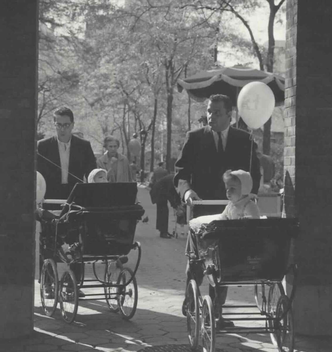 Two men in suits push vintage baby carriages with small children inside. One child holds a balloon. Several people walk in the background along a tree-lined path on a sunny day, possibly at a park or public event.