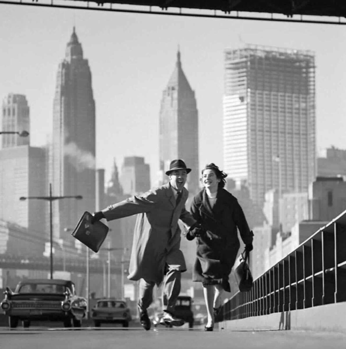 A man and woman in 1950s attire run joyfully on a city street with tall skyscrapers and vintage cars in the background, possibly New York City. The man holds a briefcase, and both look happy and energetic.
