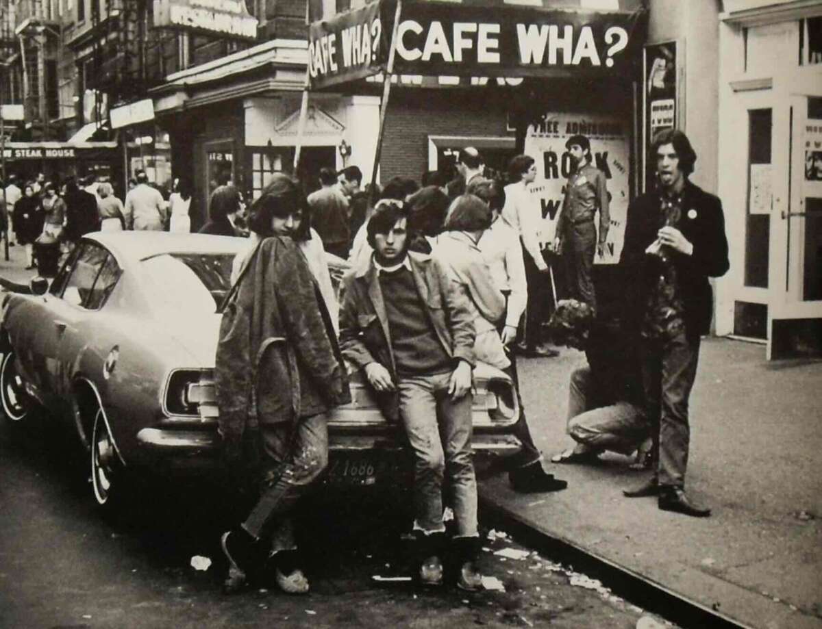 Black-and-white photo of young people gathered outside Cafe Wha? in an urban setting, some leaning on a vintage car, others standing or sitting near the entrance, evoking a 1960s or 1970s counterculture vibe.