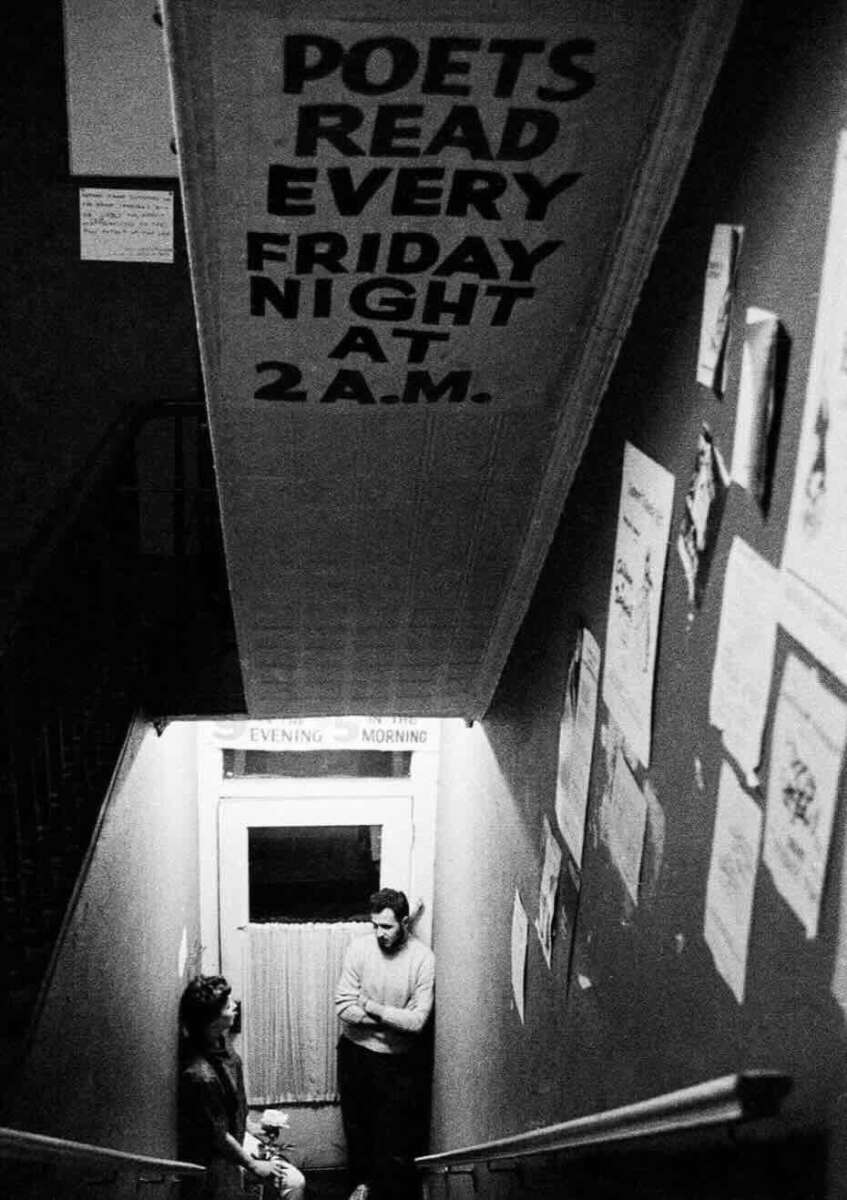 Black and white photo of two people talking at the bottom of a stairwell, with posters on the walls. A sign above reads, "POETS READ EVERY FRIDAY NIGHT AT 2 A.M.