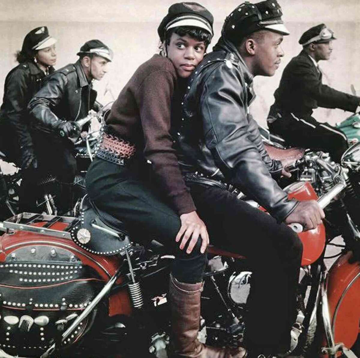 A group of Black men and a woman in leather jackets and hats sit on motorcycles, with the woman sitting sideways behind one of the riders, looking toward the camera. The scene appears vintage and vibrant.