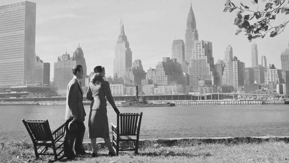 A man and woman in formal 1940s attire stand by two empty benches, looking across a river at the Manhattan skyline with iconic skyscrapers, including the Empire State and Chrysler buildings, on a clear day.