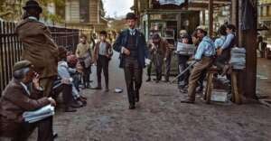 A colorized historical street scene shows men and boys in early 20th-century attire, some selling newspapers, as a man in a suit and bowler hat walks by; buildings and a newsstand are visible in the background.