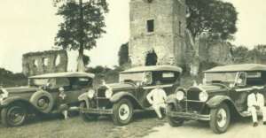 Three vintage cars with people sitting on or near them are parked in front of an old stone ruin surrounded by trees. The image appears to be from the early 20th century.