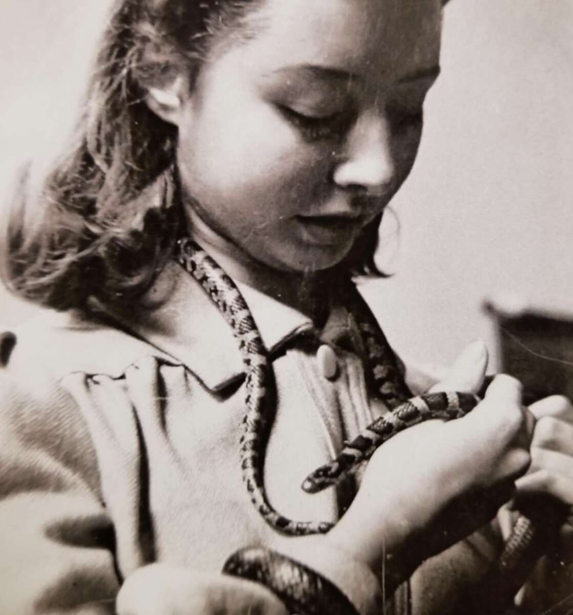 A young woman with wavy hair looks down at a snake draped around her neck and arm, gently holding it in her hands. The image is in black and white.