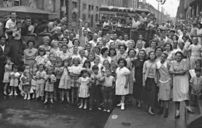 A large crowd of men, women, and children gather on a city street for a group photo. The image appears to be from the mid-20th century, with people dressed in vintage clothing, smiling and looking toward the camera.