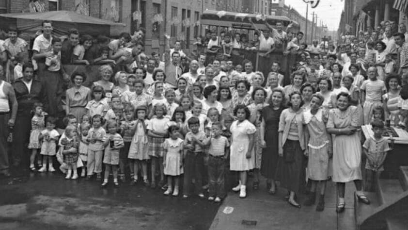 A large crowd of men, women, and children gather on a city street for a group photo. The image appears to be from the mid-20th century, with people dressed in vintage clothing, smiling and looking toward the camera.