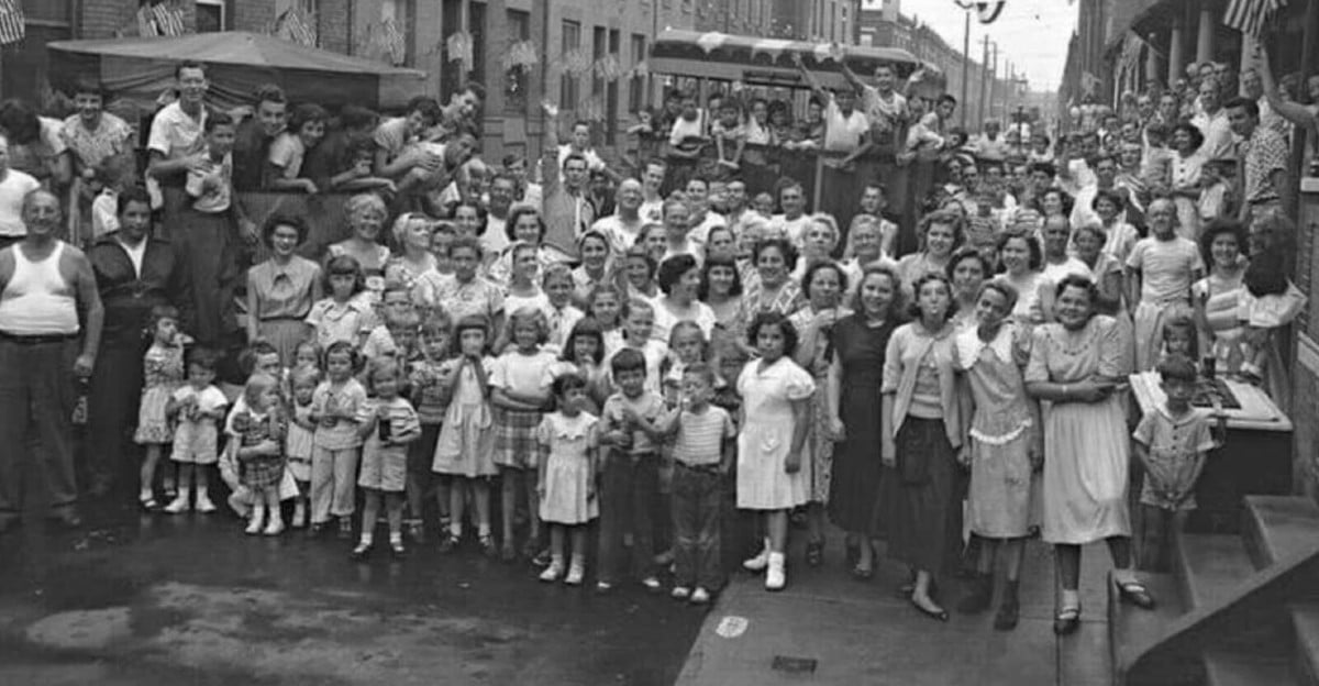 A large crowd of men, women, and children gather on a city street for a group photo. The image appears to be from the mid-20th century, with people dressed in vintage clothing, smiling and looking toward the camera.
