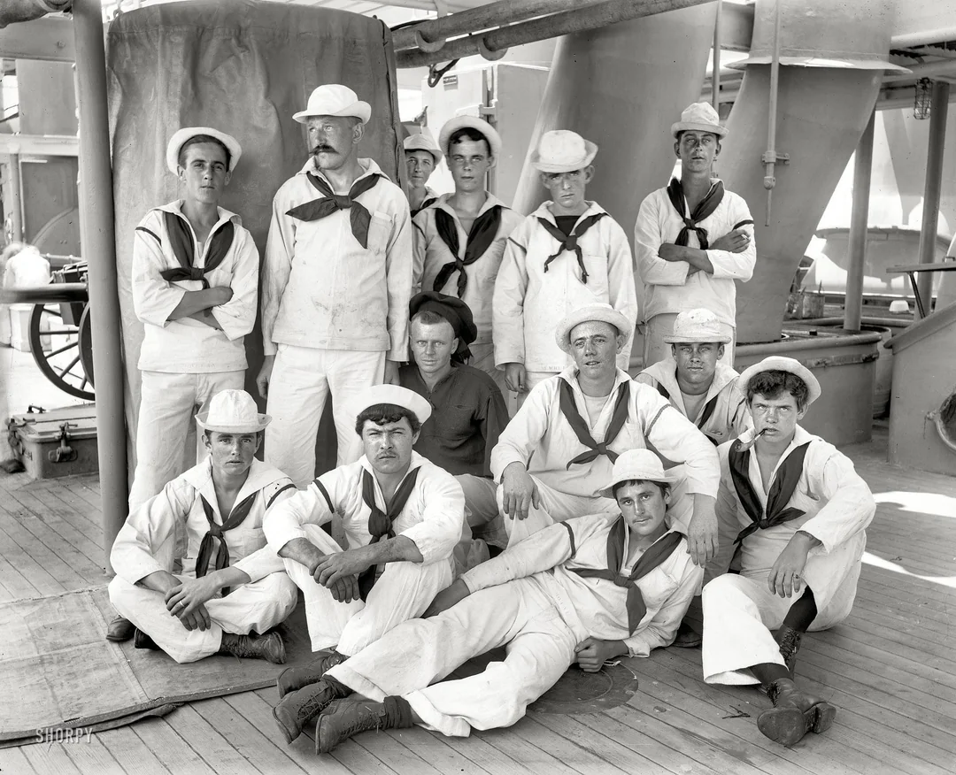 Eleven sailors in white uniforms and hats pose together on the deck of a ship, with some standing and others sitting or reclining on the wooden floor, surrounded by large equipment and pipes.