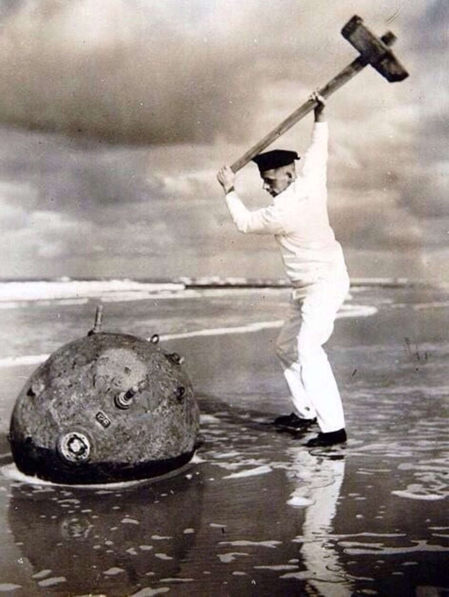 A sailor in a white uniform raises a large hammer to strike a naval mine on a beach, with the ocean and cloudy sky in the background.