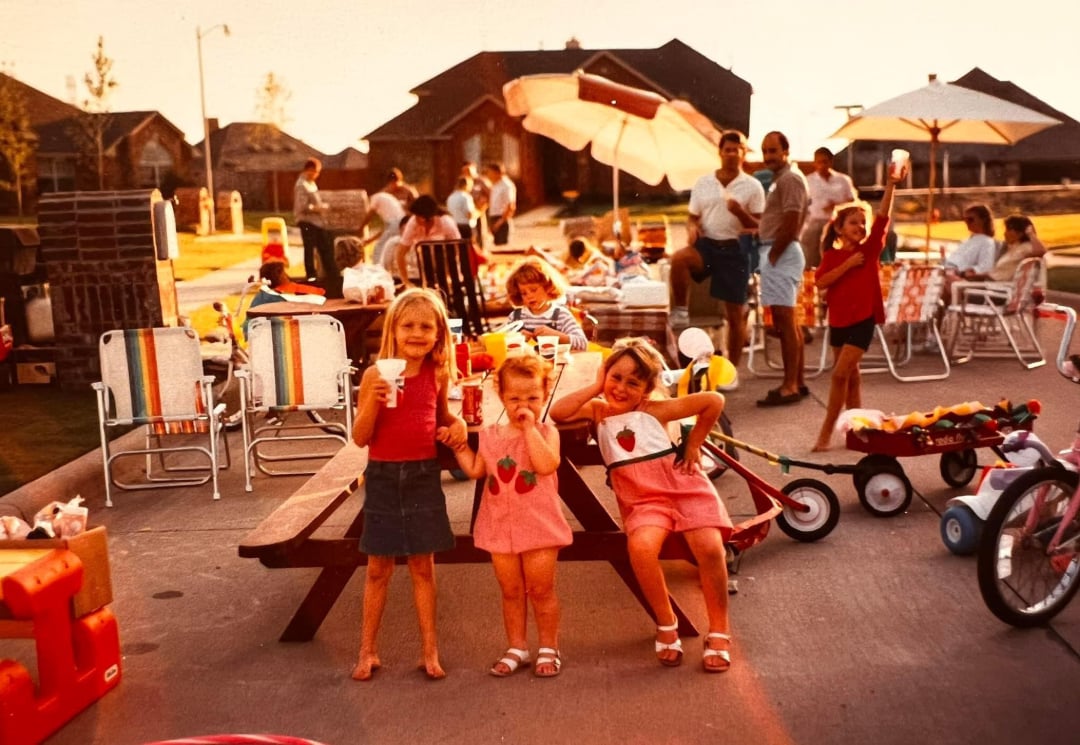 Three young girls in summer clothes stand on a picnic table, smiling and posing at a lively outdoor block party. People mingle in the background among lawn chairs, umbrellas, and children's toys on a suburban street.