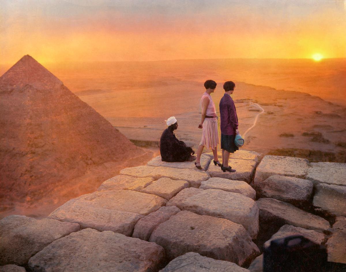 Two women and a man sit and stand on ancient stone blocks, overlooking the pyramids of Giza at sunset. The sky is orange and hazy, with the desert stretching into the distance.
