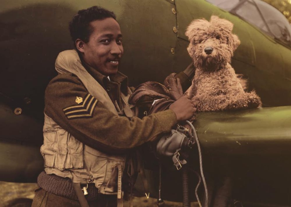 A smiling WWII-era pilot in uniform stands beside a military aircraft, holding a leather helmet. A small, fluffy dog sits on the wing next to him.