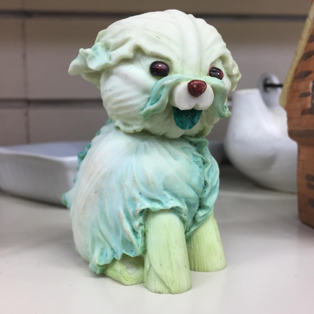 A small figurine of a dog made to look like it is sculpted from leafy cabbage, with greenish-white coloring, red nose, and dark round eyes, displayed on a shelf.