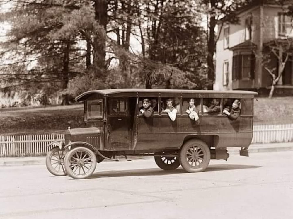A vintage school bus with several children leaning out the windows, parked on a street in front of a house and trees, in a black-and-white photo from the early 20th century.