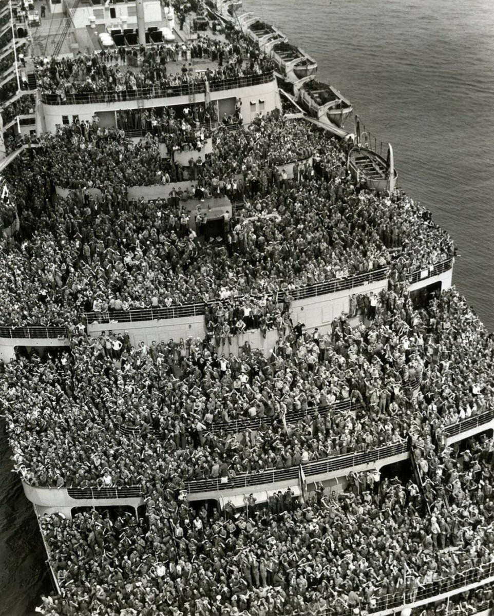 A large ship is packed with thousands of people crowded on multiple decks, many waving toward the camera. The scene appears festive or celebratory, with the ocean visible in the background.