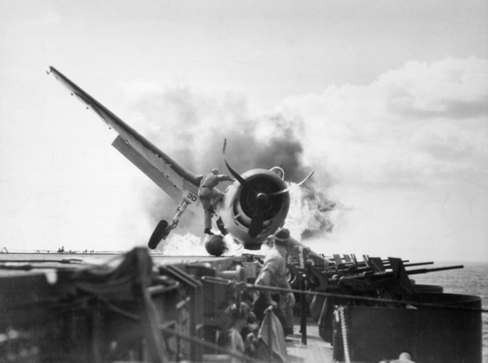 A World War II-era fighter plane crashes onto the deck of an aircraft carrier, with smoke billowing from its engine as crew members on deck look on.