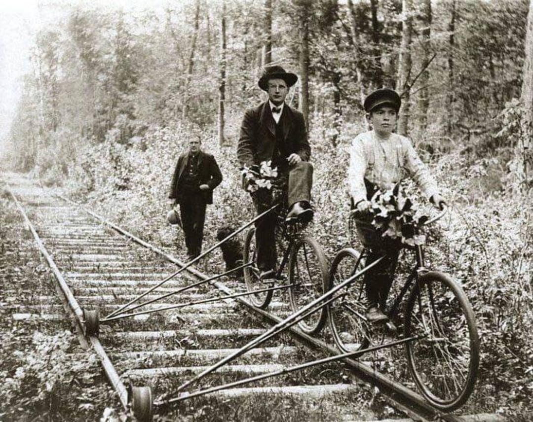 Two men ride specially modified bicycles on railroad tracks through a wooded area, while a third man walks behind them on the tracks. The bicycles have metal frames adapted to fit the rails.