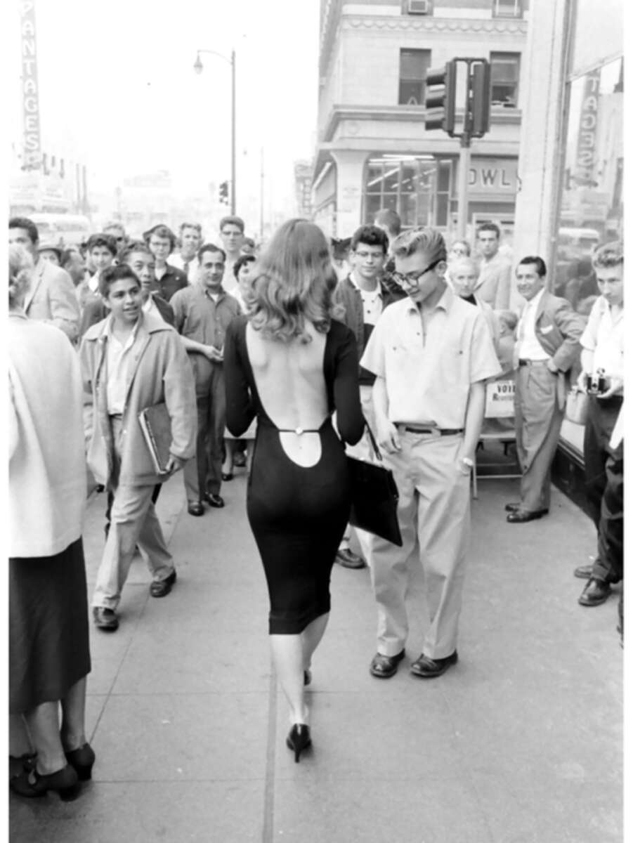 A woman in a backless black dress walks down a busy city sidewalk, drawing the attention of many men around her. Pedestrians stop and stare, and some appear surprised or amused.