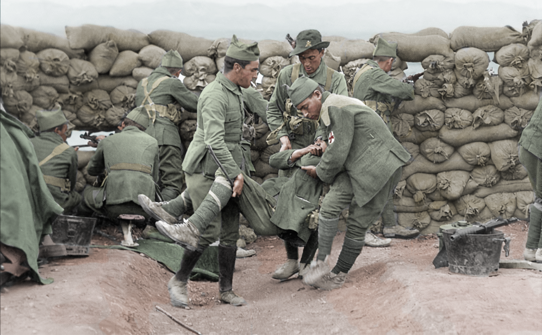 Soldiers in green uniforms tend to a wounded comrade in a trench, with sandbags stacked behind them; some soldiers offer medical assistance while others observe or assist.